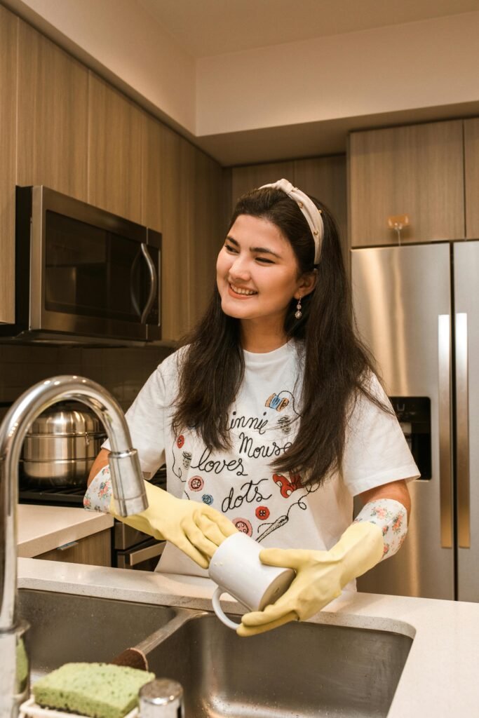 A woman in gloves washes a mug in a cozy kitchen, smiling brightly.